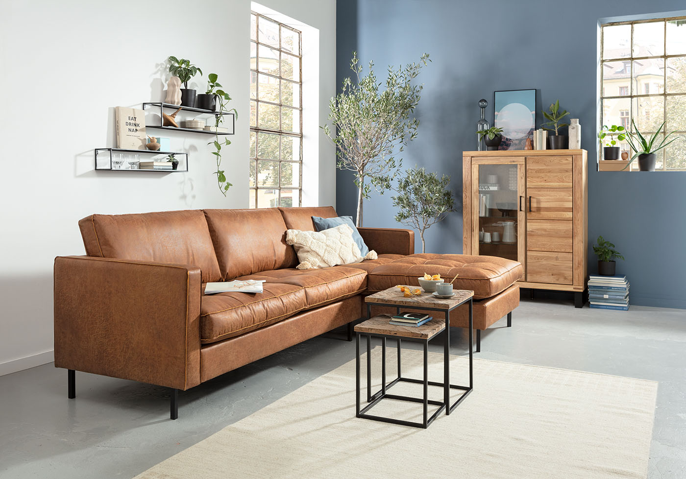 Neben einer taubenblauen Wand, die zusammen mit zwei kleinblättrigen Bäumchen an den Mediterranen Stil erinnert, steht ein Industrial-Style Sofa in freundlichem Braun im Wohnzimmer.