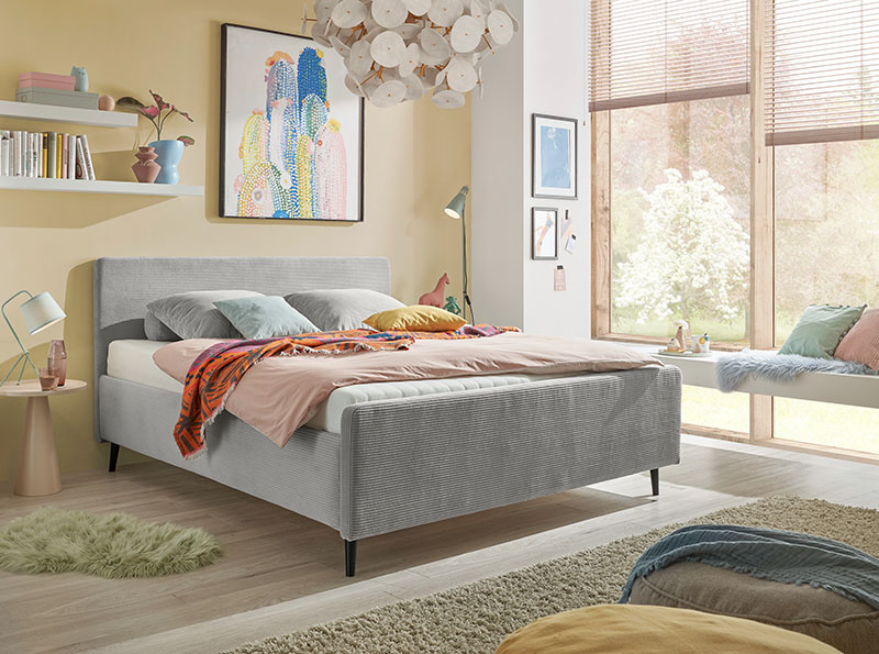Helles, freundliches Schlafzimmer mit einem grauen Polsterbett in modernem Design. Farbige Akzente durch Kissen, Decken und Wandbilder schaffen eine frische, kreative Atmosphäre. Große Fenster mit Blick ins Grüne