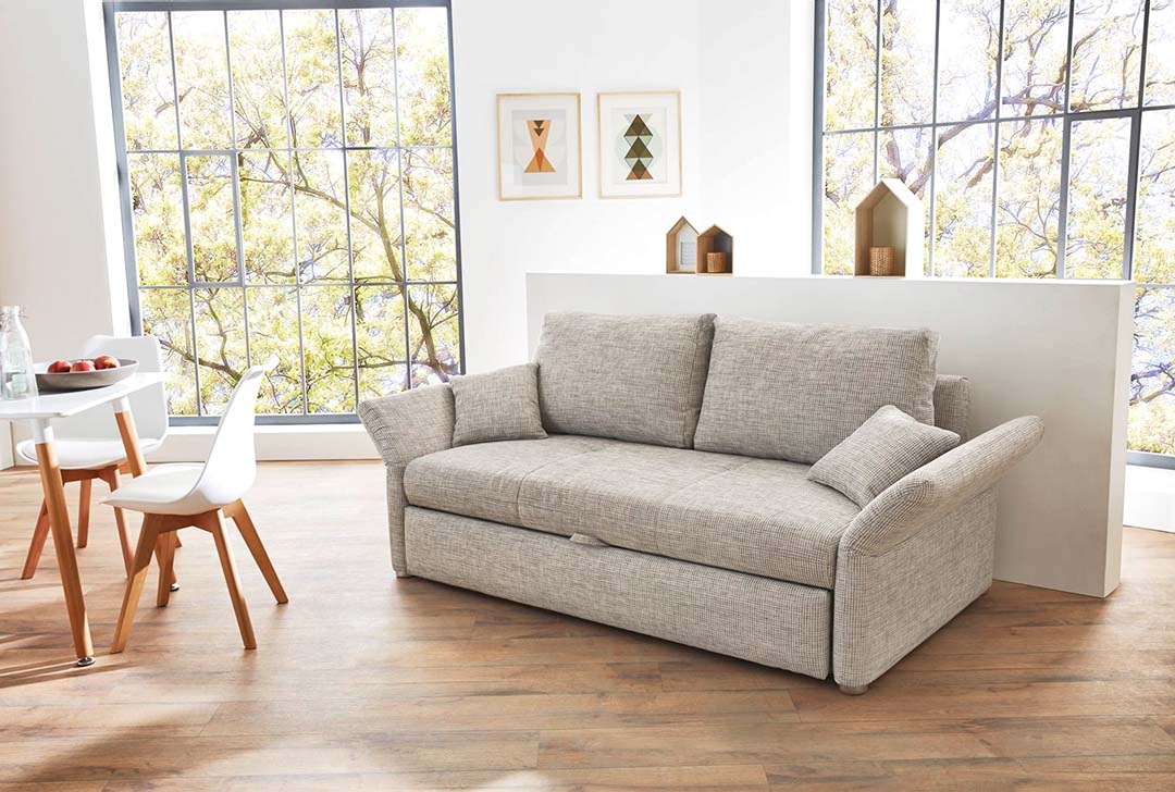Einem kleinen Wohn-Esszimmer stehen ein hellgraues Zweisitzer-Sofa und helle, einfache Stühle mit organisch geformter Sitzschale gut