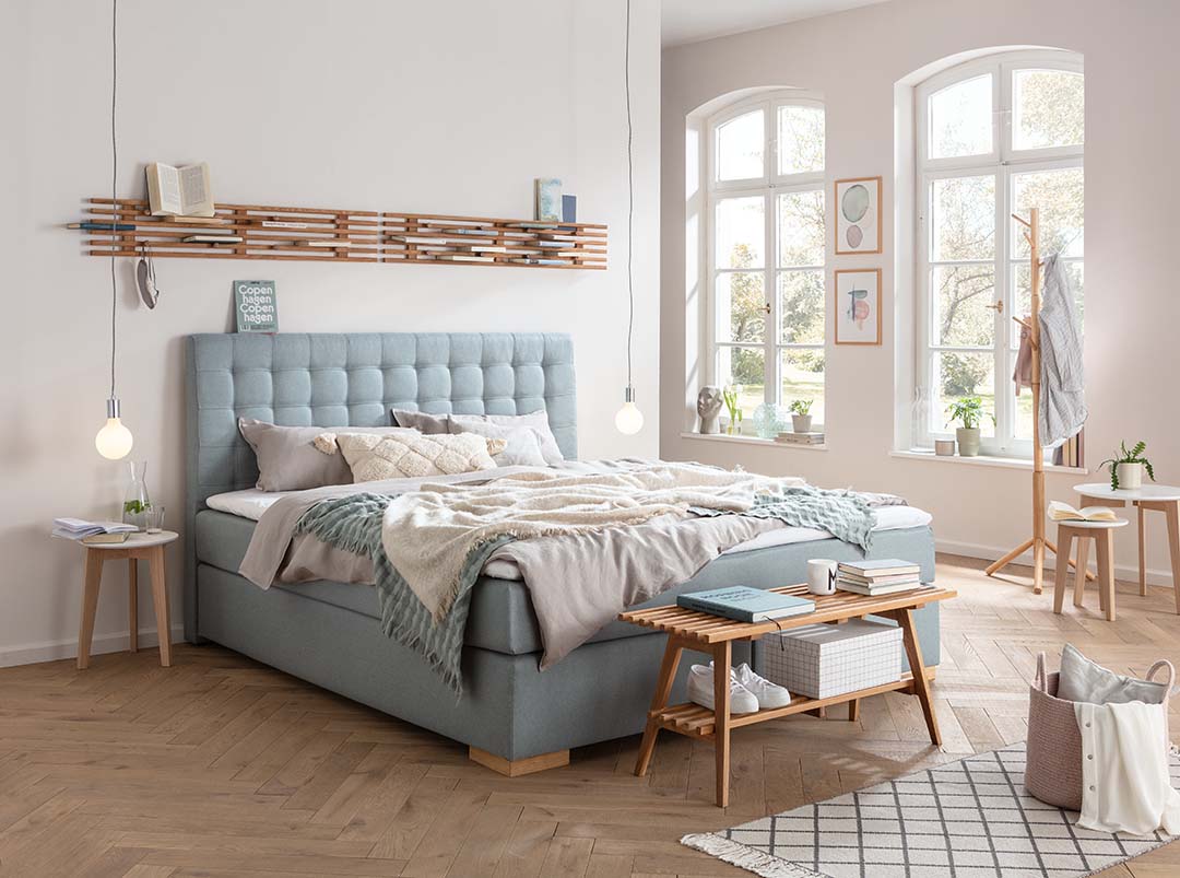 Im Schlafzimmer sind platzsparend holzfarbener Garderobenständer und Schuhbank untergebracht. Garderobenbretter sind horizontal an der Wand über dem blauen Polsterbett angebracht und erinnern durch ihre Lücken optisch an ein Floß