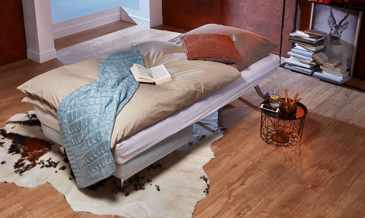 Ein Kunskuhtfell sowie ein Waldtiergemälde mit Geweihdarstellung passen wunderbar in Chalet-Stil Wohnzimmer. Dein geöffnetes Schlafsofa kannst Du dort mit Bettdecke, Wollplaid und feuerfarbenem Kissen dekorieren