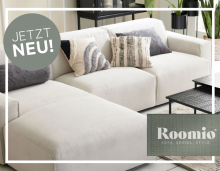 Helles, modernes Sofa mit dekorativen Kissen; links „Jetzt neu!“ Hinweis, unten Roomio Logo.