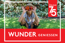 Kind mit Strohhut hockt im Garten und hält zwei Tomaten vor die Augen, daneben roter Jubiläumsbanner mit „75 Jahre“ und Schriftzug „Wunder geniessen“.  