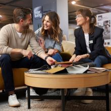 Paar sitzt auf einem Sofa in einem Möbelhaus und lässt sich von einer Interliving-Beraterin mit Stoffmustern freundlich beraten.