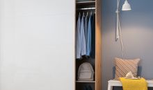 In einem Schrank mit leicht geöffneter Schiebetür sind hängende Oberteile und ein Rucksack zu sehen.
