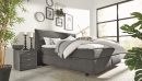 Modernes Boxspringbett mit grauem Stoffbezug, weißer Bettdecke, Kissen und Tagesdecke im Schlafzimmer.
