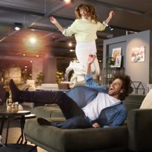 Lachendes Paar tobt ausgelassen auf einem Sofa in einer Möbelausstellung, die Frau springt freudig hinter dem Mann.