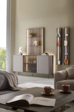 Gemütlicher Wohnraum mit Sideboard, offenem Wandregal und Wandpaneel mit Glasvitrine; im Vordergrund ein Couchtisch mit geöffnetem Buch und zwei Tassen.