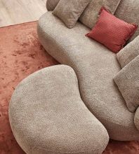 Ein hellbraunes Sofa mit organischen Formen und optisch passendem Hocker steht im Wohnzimmer, auf einem terracottafarbenen Teppich platziert und mit einem weinroten Kissen dekoriert.
