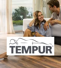 TEMPUR Wohnambiente mit Wohlfühlmoment Frau sitzt mit Tasse auf dem Boden, Mann lehnt sich zu ihr; vorn das TEMPUR Logo, im Hintergrund grosse Fenster mit Naturblick.