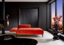 Modernes Wohnzimmer mit oranger Samtliege, schwarzer Wand, Kamin, großen Fenstern und Stadtblick bei Sonnenuntergang.