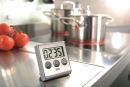 Digitaler Küchentimer auf Edelstahl-Arbeitsfläche, im Hintergrund Töpfe auf Herd und frische Tomaten.