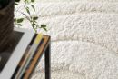 Weißer, flauschiger Teppich mit Struktur, daneben ein Tisch mit Büchern und grünem Pflanzenblatt.