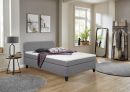 Graues Boxspringbett mit weißer Matratze, lila Decke und Kissen, modernes Schlafzimmermöbel, komfortabel.