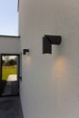 Moderne schwarze Außenwandleuchte an weißer Hauswand, leuchtet bei Tageslicht, minimalistisches Design.