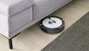 Saugroboter reinigt hellen Holzboden unter grauem Sofa neben Teppich, modernes Smart-Home-Gerät im Einsatz.