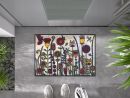 Bunte Fußmatte mit Blumenmuster vor moderner Haustür auf grauem Boden, Schuhe und Pflanzen im Bild.