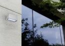 Moderne Außenwandleuchte an Betonwand neben großer Glasfassade mit Baumspiegelung und grünen Ästen