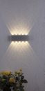 Moderne LED-Wandleuchte an weißer Wand, dekoratives Lichtmuster, gelbe Rosen im Vordergrund.