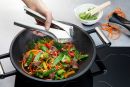 Hand presst Knoblauch in Wok mit gebratenem Gemüse, Rindfleisch und frischen Kräutern auf Induktionsherd.