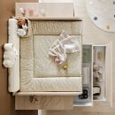Wickelkommode mit beigem Wickelauflagen, Babydecke, Spielzeug, Windeln und offenen Schubladen im Kinderzimmer.