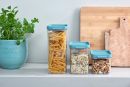Vorratsdosen mit Pasta, Müsli und Nüssen auf Küchenarbeitsplatte neben Holzbrett und Kräutertopf.