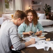 Lachendes Paar sitzt am Tisch und plant die Einrichtung mit Skizzen und Materialmustern in einem gemütlichen Wohnzimmer.