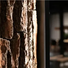 Detailaufnahme einer Wandverkleidung aus grober Baumrinde mit stimmungsvoller Beleuchtung, im Hintergrund ein unscharfer Innenraum mit modernem Ambiente.