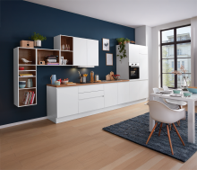 Moderne weiße Einbauküche mit Holzakzenten, Esstisch, großen Fenstern und blauer Wand in hellem Wohnraum.