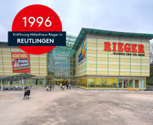 Frontansicht des Möbelhauses RIEGER in Reutlingen um 1995/1996. Das Gebäude mit gelb-grüner Fassade und großem Glasdach zeigt deutlich den roten RIEGER-Schriftzug. Eine Menschenmenge und Werbeschilder wie „Achtung! Preise gesenkt!“ beleben die Szene.
