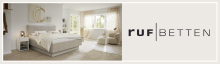 Modernes Schlafzimmer mit RUF Boxspringbett RUF Betten Logo mit beigem Boxspringbett in hellem, modernem Schlafzimmer mit Fenstern und Sitzecke.