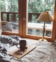 Einem Interieur im Chalet-Stil mit Blick aus dem Holzfenster verleihen Grobstrick-Plaid, Teetasse und ein beigefarbener Lampenschirm optische Wärme