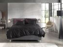 Modernes Schlafzimmer mit grauem Boxspringbett, schwarzer Bettwäsche, großen Fenstern und stilvoller Einrichtung.