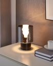 Moderne Tischlampe mit rauchgrauem Glasschirm leuchtet auf weißem Nachttisch neben Büchern und Kaffeetasse.