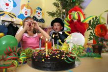 „Kindergeburtstag mit bunt verkleideten Kindern, Luftschlangen, Ballons und einem Schokoladenkuchen mit Kerzen – fröhliche Partydekoration und Wandbilder mit Clown-Motiven im Hintergrund.