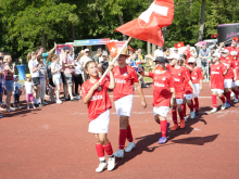 Kindertanzgruppe in rot-weisser Kleidung mit Fahne bei einer Veranstaltung im Freien.