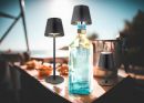 Drei moderne Tischlampen und eine Glasflasche auf einem Holztisch im Sonnenlicht, Outdoor-Szene.