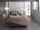 Modernes Schlafzimmer mit grauem Boxspringbett, brauner Bettwäsche, großen Fenstern und minimalistischer Einrichtung.