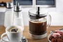 Französische Kaffeepresse mit frisch gebrühtem Kaffee, Tasse, Milch, Zucker und Gebäck auf Holztisch.