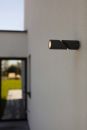 Moderne schwarze Außenwandleuchte an weißer Hauswand, beleuchtet, minimalistisches Design, Tageslicht.