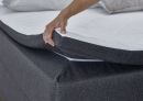Hand zieht weißen Matratzentopper auf graues Boxspringbett, Bettwäsche und Kissen im Hintergrund, Schlafzimmer.