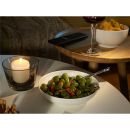 Schale mit grünen Oliven und Chili, Kerze, Chips, Rotweinglas und Fernbedienung auf Couchtisch bei Abendstimmung.