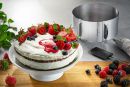 Sahnetorte mit frischen Beeren und Minze auf Tortenplatte, Backzubehör und Beeren im Hintergrund, Food-Fotografie.