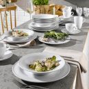 Weißes Porzellangeschirr mit Salat und Gemüse auf modern gedecktem Esstisch, frische Kräuter im Hintergrund.