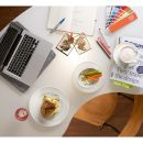 Moderner Arbeitsplatz mit Laptop, Notizbuch, Farbfächer, Kaffee, Sandwich und Gemüsesticks mit Dip.