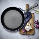 Antihaft-Pfanne auf Holzbrett mit Knoblauch, roten Zwiebeln, Frühlingszwiebeln und Küchentuch, Küchenzubehör