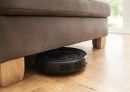 Saugroboter reinigt Holzboden unter braunem Sofa im Wohnzimmer bei Tageslicht, smartes Haushaltsgerät.