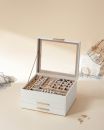 Elegante Schmuckbox aus weißem Leder mit Glasdeckel, goldenen Griffen und Fächern zur Schmuckaufbewahrung.