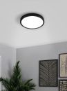 Moderne LED-Deckenleuchte rund, schwarz, leuchtet in hellem Raum mit Pflanze und abstrakter Wandkunst.