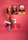 Frau mit blondem Bob, neonpinkem Brillenrahmen und gelber Retro-Mode vor rotem, mandalaartigem Hintergrund.